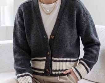 May include: A charcoal grey knit cardigan featuring a V-neckline, long sleeves, and a button-down front. The garment includes thick cream-coloured ribbed cuffs and a matching cream-coloured ribbed hem, both accented with thin charcoal grey stripes.