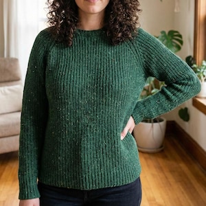 May include: A green knit sweater with a ribbed texture. The long-sleeved sweater has a crew neck and a speckled pattern. The model is wearing the sweater with their hand on their hip. The sweater is made of a soft, comfortable material.