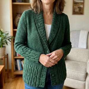 May include: A dark green knitted cardigan with a shawl collar and button closure. The cardigan features cable knit detailing on the sleeves and front panels. The person is wearing a white top underneath and blue jeans.