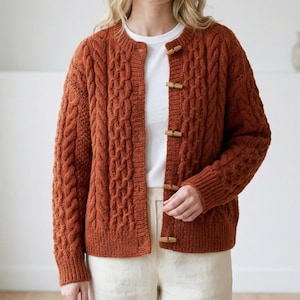May include: A rust-colored cable-knit cardigan with wooden toggle closures. The sweater features a round neckline and a textured pattern. The sleeves are long, and the cardigan appears to be made of a soft, warm material.