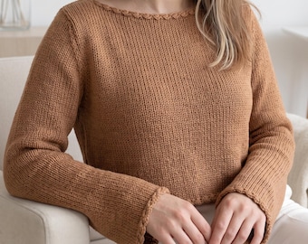 May include: A long-sleeved, tan-coloured knitted jumper featuring a boat neckline and matching braided trim detailing along the collar and sleeve cuffs. The garment is constructed with a standard stockinette stitch pattern throughout the body.