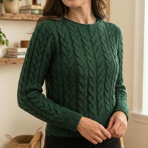 May include: A dark green cable-knit jumper with a crew neck and long sleeves. The jumper has a textured, woven pattern across the body and sleeves. The garment is made of a soft material, possibly wool.