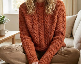 May include: A burnt orange cable-knit jumper with a round neck. The jumper has a textured design on the sleeves and front. The person is wearing brown corduroy trousers. The background includes a window, a table, and a sofa.