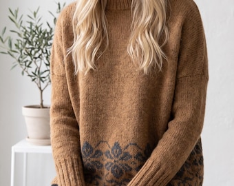 May include: A long-sleeved, crew-neck jumper in a warm tan colour, featuring a dark grey floral pattern along the bottom hem. The garment has a ribbed knit collar, cuffs, and hem, with a relaxed fit and a textured, soft-looking knit construction.