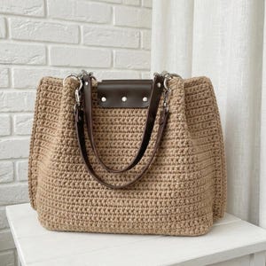 May include: A beige crochet tote bag with brown leather handles and accents. The bag features a rectangular shape with a flap closure and silver-tone hardware. The bag is sitting on a white surface.