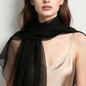 May include: A sheer black scarf draped around the neck and shoulders. The scarf is made of a lightweight, flowing fabric. The model is wearing a beige satin camisole. The background is a plain white.