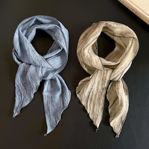 May include: Two silk scarves, one in a muted blue and the other in a beige tone, are displayed on a dark surface. Both scarves have a crinkled texture and are tied in a loose knot, with the ends flowing freely.