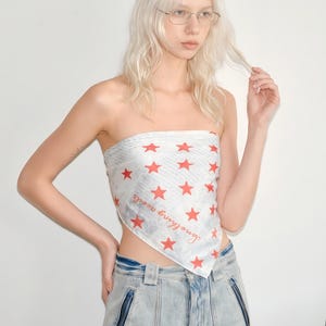 Puede incluir: Top bandana blanco con estampado de estrellas rojas y la frase "Something needs". Se combina con vaqueros de mezclilla de lavado claro. El estilo general es casual y juvenil.