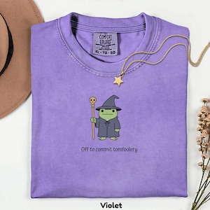 Peut inclure: Un t-shirt violet Comfort Colors avec une grenouille de dessin animé habillée en magicien, tenant un bâton. Le texte "Off to commit tomfoolery" est en dessous. Un collier avec un pendentif étoile doré et un chapeau marron sont visibles.