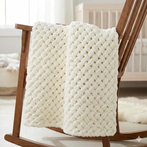 Puede incluir: Una manta de punto grueso de color crema sobre una mecedora de madera. La manta presenta un patrón tejido y una textura suave y afelpada. La silla está en un entorno de guardería, lo que sugiere un artículo acogedor y cómodo para un bebé o un niño.