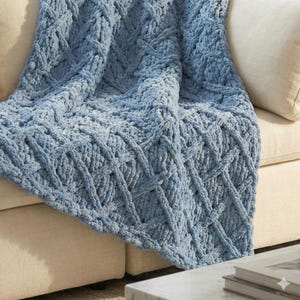 Op de afbeelding: Een zachte, blauwe gebreide sprei met een textuur, ruitvormig ontwerp. De sprei is over een beige bank gedrapeerd en toont zijn zachte, gezellige uitstraling. De afmetingen van de sprei zijn niet zichtbaar.