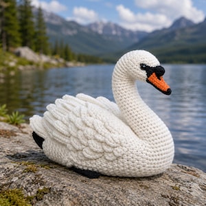 May include: A handmade white crochet swan with a curved neck and orange beak. The swan has black accents on the wing tips and beak. The swan is sitting on a rock with a lake and mountains in the background.