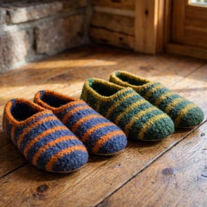 May include: Two pairs of wool slippers. One pair is navy blue with orange stripes, and the other is green with yellow stripes. The slippers are on a wooden surface, with a brick wall and window in the background.