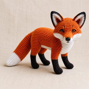 Könnte beinhalten: Ein gehäkeltes Fuchskuscheltier mit leuchtend orangefarbenem Körper, weißer Brust und Schnauze sowie schwarzen Unterbeinen. Der Fuchs hat spitze Ohren mit weißen Innendetails, dunkle Augen, eine schwarze Nase und einen langen, buschigen Schwanz mit weißer Spitze.