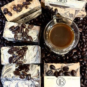 May include: Several bars of Misty Bubbles Coffee Soap are arranged with coffee beans and a glass of coffee. The soap bars are rectangular, with a marbled design and topped with coffee beans. The soap packaging includes the text "Misty Bubbles Coffee Soap".