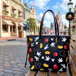 May include: Black tote bag with a pattern of Mickey Mouse gloves, shoes, and face outlines in white, yellow, and red. The bag has black handles with red trim. The bag is on a wooden bench.