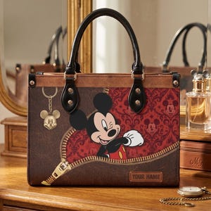 May include: Brown tote bag featuring Mickey Mouse peeking out from a zipper design. The bag has a red and black damask pattern, a gold Mickey Mouse charm, and black handles. The text "YOUR NAME" is visible on the bag.