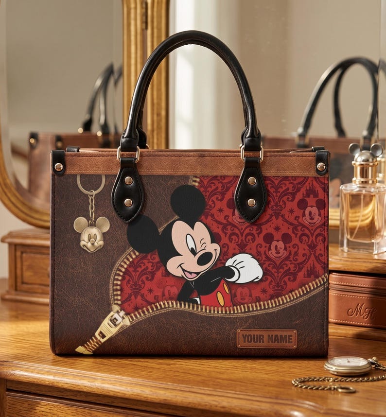 May include: Brown tote bag featuring Mickey Mouse peeking out from a zipper design. The bag has a red and black damask pattern, a gold Mickey Mouse charm, and black handles. The text "YOUR NAME" is visible on the bag.