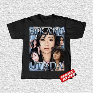 May include: Black t-shirt featuring a graphic of Hikaru Utada with the text "HIKARU UTADA" in a stylized font. The design includes multiple images of the artist with a lightning bolt effect. The shirt has a tag that says "KAOS 1026".