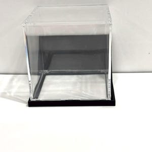 May include: A clear acrylic display box with a black base. The box is cube-shaped with a transparent top and sides, showcasing the contents within. The black base provides a contrasting foundation for the clear structure.