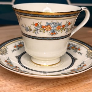 May include: A white teacup and saucer set with a floral design. The cup and saucer have a cream-colored base with a gold rim and a blue and gold border. The floral design includes orange, blue, and yellow flowers.
