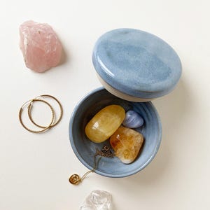May include: A light blue ceramic trinket box with lid, open to reveal crystals and a gold necklace. Rose quartz, gold hoop earrings, and other crystals are scattered around the box. The crystals are various colors, including yellow, orange, and clear.