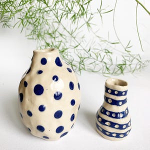 Può includere: Due vasi in ceramica. Il vaso più grande è beige con pois blu navy. Il vaso più piccolo è beige con strisce e punti orizzontali blu navy. Entrambi i vasi hanno un aspetto rustico e fatto a mano.