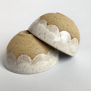Può includere: Due piccole ciotole in ceramica rotonde con una base in argilla beige maculata e uno smalto bianco lucido che copre la parte inferiore. Lo smalto bianco presenta un design a bordo smerlato che rivela l'argilla beige naturale e testurizzata.