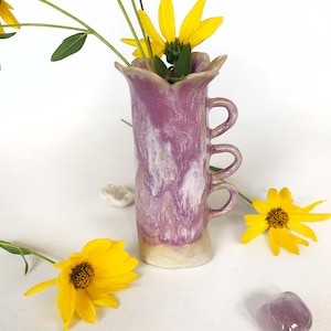 Può includere: Un vaso in ceramica con smalto marmorizzato viola e bianco, caratterizzato da un bordo smerlato e tre manici circolari sovrapposti su un lato. Il vaso contiene fiori gialli con steli e foglie verdi, accompagnato da una piccola pietra viola.