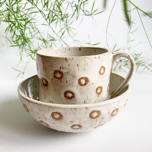Könnte beinhalten: Ein Keramik-Tassen- und Schüssel-Set. Die cremefarbene Keramik weist braune kreisförmige Designs auf. Die Tasse steht in der Schüssel. Das Set steht auf einer weißen Oberfläche, mit grünem Laub im Hintergrund.