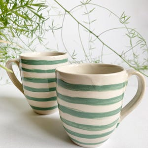 Può includere: Due tazze in ceramica con base color crema e strisce orizzontali verdi. Le tazze hanno una forma arrotondata con manico e sono poste su uno sfondo bianco con fogliame verde.