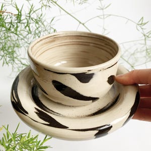 Könnte beinhalten: Cremefarbenes Keramikschalen- und Untertassen-Set mit dunkelbraunen Pinselstrich-Designs. Die Schale hat eine runde Form mit leicht strukturierter Oberfläche. Die Untertasse ist kreisförmig, mit einem erhöhten Rand und passenden Pinselstrichen.