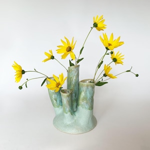 Könnte beinhalten: Eine mehrteilige Keramikvase mit einer texturierten, blass minzgrünen Glasur und bläulichen Akzenten. Die Vase verfügt über vier verschiedene zylindrische Öffnungen unterschiedlicher Höhe, die jeweils eine leuchtend gelbe Blume mit brauner Mitte halten.