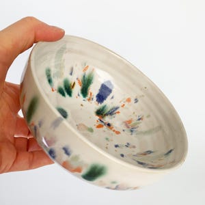 Könnte beinhalten: Eine weiße Keramikschale mit einem gesprenkelten Design. Die Schale hat Farbspritzer in Grün, Blau und Orange. Die Schale wird von einer Hand vor einem weißen Hintergrund gehalten.
