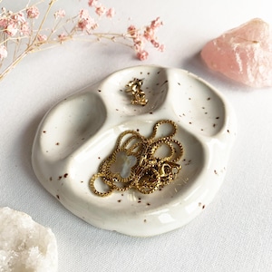 Pode incluir: Um prato de cerâmica branca com três compartimentos arredondados e manchas castanhas. O prato contém um colar dourado com um pendente em forma de trevo e um pequeno brinco dourado. A superfície é brilhante e tem uma forma irregular.