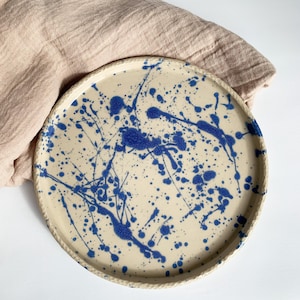 Può includere: Un piatto rotondo in ceramica con base color crema e un design a spruzzi blu. Il piatto ha un bordo strutturato a forma di corda. Il piatto è appoggiato su un panno beige.