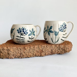 Puede incluir: Dos tazas de cerámica con diseños florales azules. Una taza es redonda, la otra cilíndrica. Ambas tazas tienen una base de color crema y están decoradas con flores y tallos azules. Se encuentran sobre un ladrillo marrón rojizo texturizado.