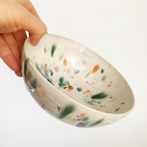 Può includere: Una ciotola in ceramica fatta a mano con un design maculato. La ciotola bianco sporco presenta spruzzi colorati di arancione, verde, blu e viola. La ciotola è tenuta ad angolo, mostrando l'interno e il design. Un pezzo unico per servire o esporre.