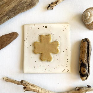 Könnte beinhalten: Eine quadratische Keramikfliese mit glänzender cremeweißer Oberfläche und braunen Sprenkeln. In der Mitte befindet sich ein erhabenes, glattes, goldbraunes vierblättriges Kleeblatt. Ein kleines kreisförmiges Loch befindet sich oben mittig.