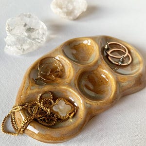 Könnte beinhalten: Eine ovale, beigefarbene Keramikschale mit fünf kreisförmigen Vertiefungen. Die Schale hat eine glänzende Oberfläche und enthält goldfarbenen Schmuck, darunter eine Kette mit einem kleeblattförmigen Anhänger und zwei kleine Creolen.