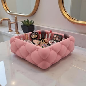 May include: A square, light pink storage tray featuring a tufted, button-detailed exterior texture. The tray is filled with various items including makeup brushes, lipsticks, a small eyeshadow palette, and several pieces of gold-toned jewellery.