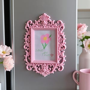 Puede incluir: Un marco de fotos rosa adornado con un dibujo infantil de una flor y las palabras "Love you Mum". El marco tiene un diseño decorativo de volutas. Una taza rosa y un jarrón con flores están en el fondo, sobre una superficie gris.