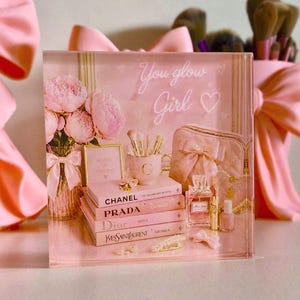 May include: A pink decorative display featuring a stack of fashion books, makeup brushes, perfume, and a cosmetic bag. The text "You glow Girl" is written on the display. A vase of pink flowers and a gold framed picture are also present.