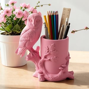 Op de afbeelding: Een roze bureau-organizer in de vorm van een uil met een takbasis. De organizer bevat potloden, pennen en een houten liniaal. Een plant in een pot met roze bloemen staat op de achtergrond. De bureau-organizer is een decoratief item.