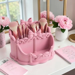 May include: A pink makeup brush organiser with a bow design, holding various makeup brushes. The organiser is on a white surface, with pink peonies in the background. A mirror and notebooks are also visible.