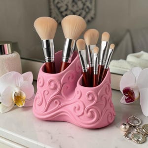 May include: A light pink makeup brush holder with a raised swirling scroll pattern, featuring two distinct compartments. The holder contains seven makeup brushes with silver metal ferrules, dark brown handles, and soft, light-coloured bristles.