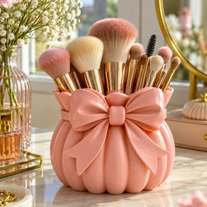 Op de afbeelding: Een lichtroze make-up kwastenhouder in de vorm van een afgerond tasje met een grote, opvallende strik aan de voorzijde. De houder is gevuld met diverse make-up kwasten voorzien van goudkleurige hulzen en zachte, veelkleurige synthetische haren.