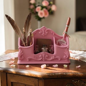 Puede incluir: Un organizador de escritorio rosa con detalles ornamentados, que contiene plumas, bolígrafos y pequeños frascos de tinta. El organizador tiene múltiples compartimentos y un diseño decorativo, perfecto para una estética vintage o romántica. El organizador está sobre una mesa de madera.