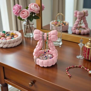 Puede incluir: Un organizador de joyas de color rosa claro con una base redondeada y un soporte vertical en forma de lazo grande. El organizador está construido con segmentos de tela acolchada y contiene varios collares y anillos en su bandeja circular.