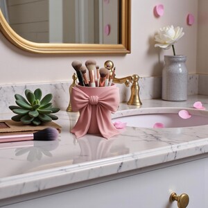 May include: A pink makeup brush holder shaped like a bow, filled with various makeup brushes. The holder sits on a marble countertop next to a succulent and a makeup brush. A gold-framed mirror and faucet are in the background.